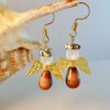 Beaded angel earrings with golden wings and brown teardrop bodies, hanging on gold hooks, displayed against a seashell.