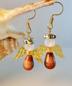 Beaded angel earrings with golden wings and brown teardrop bodies, hanging on gold hooks, displayed against a seashell.