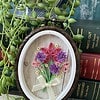 Quilled flower art in an oval frame, with pink and purple flowers, a yellow ribbon, surrounded by green vines and stacked books.