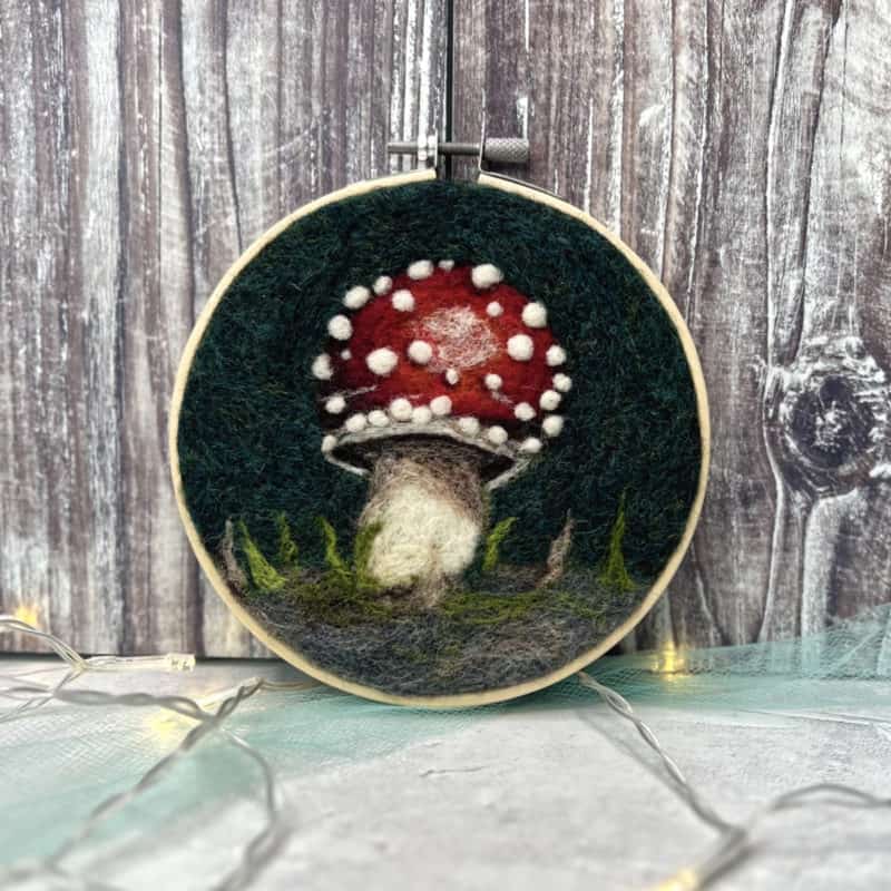 Felted mushroom art inside a wooden embroidery hoop, against a wooden background with string lights on the surface.