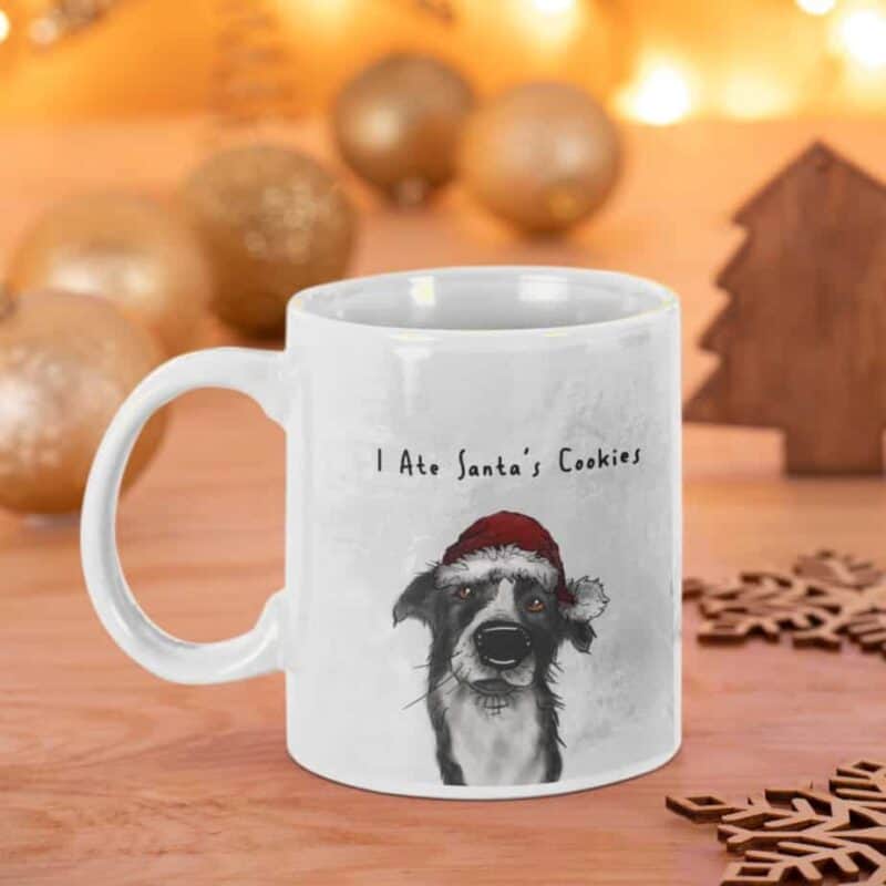 White mug with a dog wearing a Santa hat and text "I Ate Santa's Cookies." Festive background with ornaments and wooden decor.