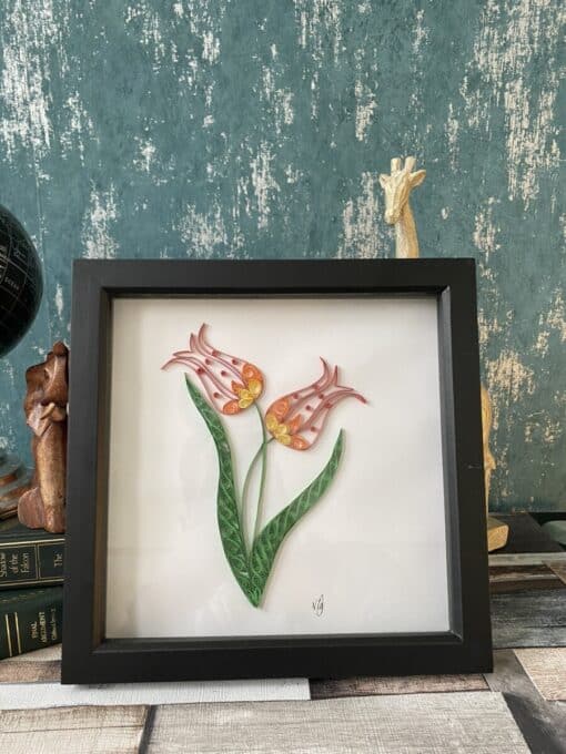 Framed quilling art features two pink tulip flowers with green leaves, placed on a rustic wood surface beside books and ornaments.