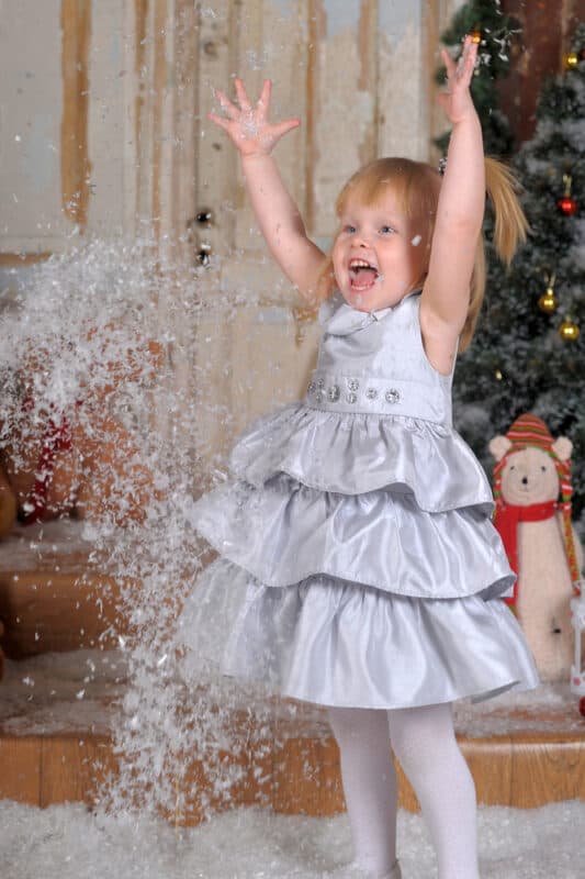 A young girl in a silver dress joyfully plays with artificial snow indoors, with a decorated Christmas tree in the background.