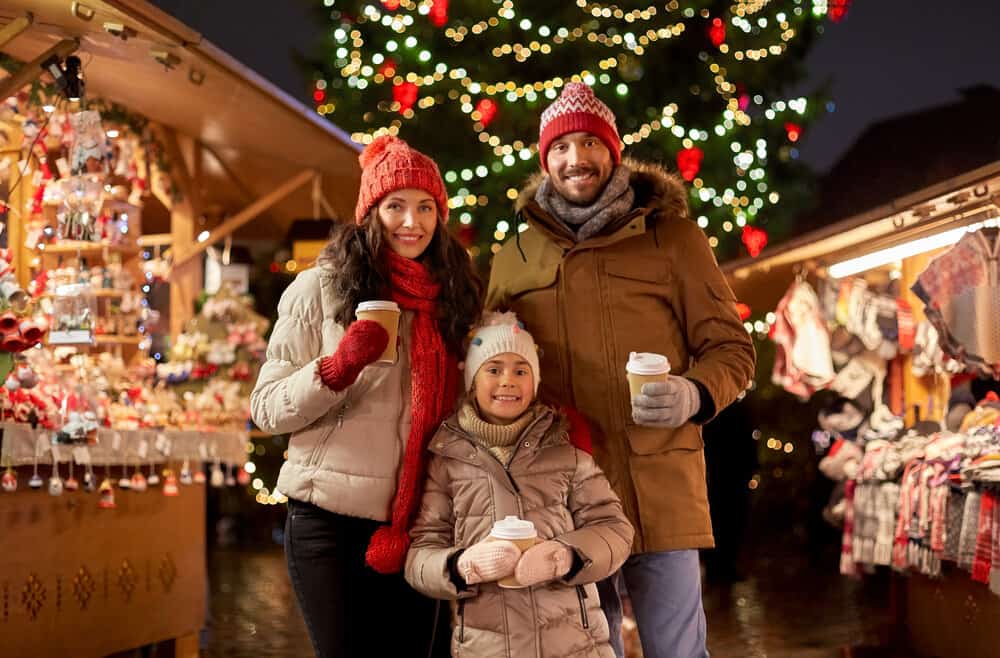 Family dressed warmly enjoying a Christmas market at night, holding hot drinks with a decorated Christmas tree in the background.