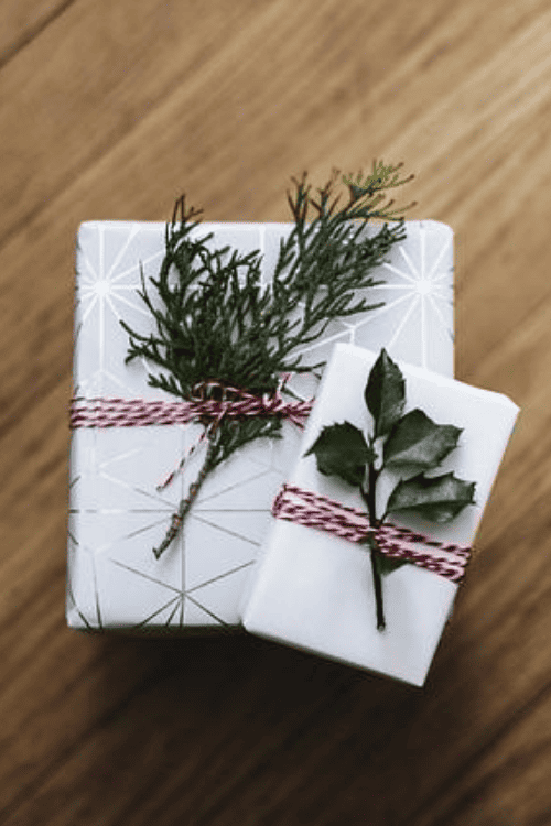 Two wrapped gifts on a wooden surface, decorated with red and white twine and green sprigs of foliage.