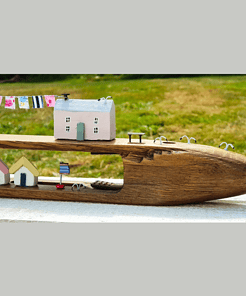 A wooden carving of a boat features miniature houses, clotheslines, and a small flag, set against a grassy background.