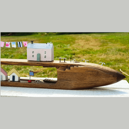 A wooden carving of a boat features miniature houses, clotheslines, and a small flag, set against a grassy background.