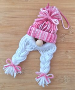 Yarn gnome ornament with a pink-striped hat, white braided yarn hair, and pink bows, lying on a wooden surface.