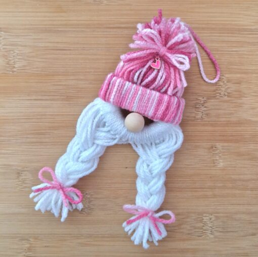 Yarn gnome ornament with a pink-striped hat, white braided yarn hair, and pink bows, lying on a wooden surface.