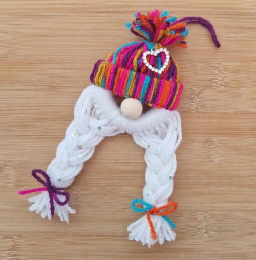 Handmade yarn gnome with a rainbow hat, white braided beard, and colorful bows, on a wooden surface.