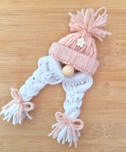 Knitted decoration with a pink hat, white braids adorned with beads, wooden nose, and pink tassel bows on a wooden surface.