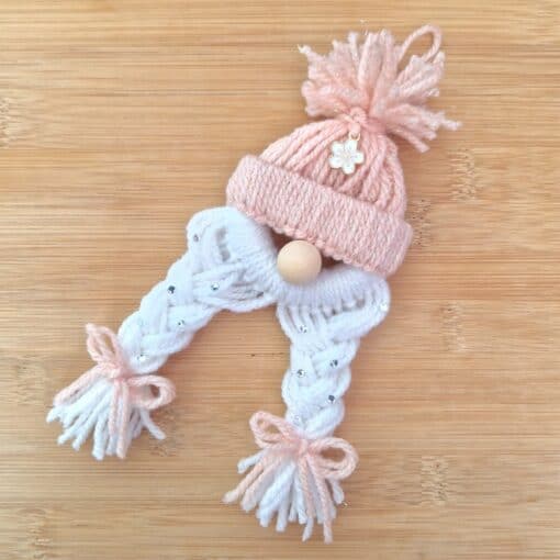 Knitted decoration with a pink hat, white braids adorned with beads, wooden nose, and pink tassel bows on a wooden surface.