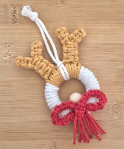 Macrame reindeer ornament with brown antlers, white face, red nose, and red bow against a wooden background.