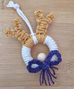 Handmade macramé reindeer ornament with antlers, a white ring body, a wooden bead nose, and a purple bow, on a wooden surface.