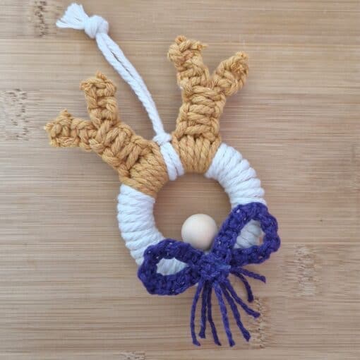 Handmade macramé reindeer ornament with antlers, a white ring body, a wooden bead nose, and a purple bow, on a wooden surface.