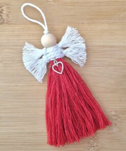 Handmade angel ornament with white wings, red tassel body, and a heart charm, lying on a wooden surface.