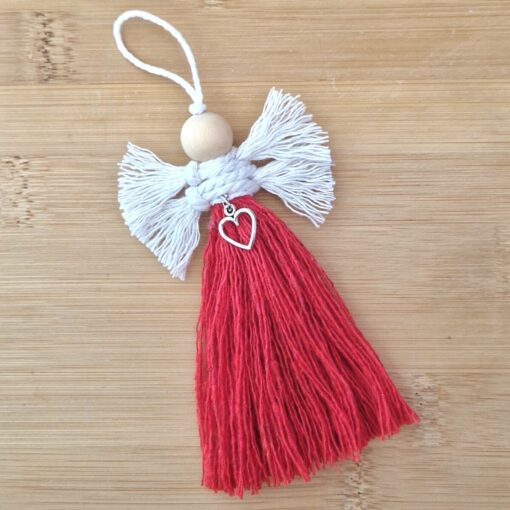 Handmade angel ornament with white wings, red tassel body, and a heart charm, lying on a wooden surface.