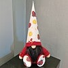 Small gnome figurine in red clothes, with a tall conical hat and shoes decorated with ladybug designs, sits on a black surface.