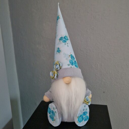 A small gnome figurine with a white beard wears a tall, pointy hat adorned with blue floral patterns and sits on a black surface.