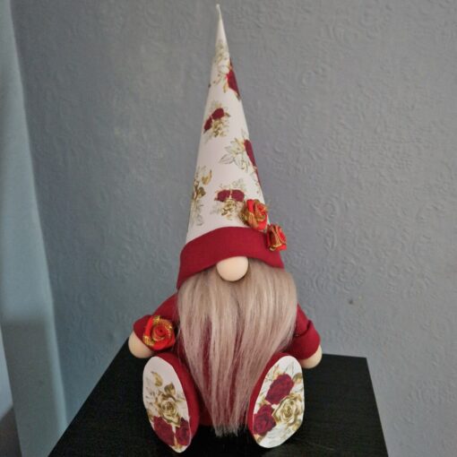 A gnome doll with a long white beard, wearing a floral hat and red clothing, sits on a black surface with a textured wall background.