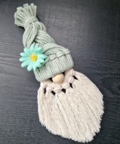 Macrame gnome with braided hat, wooden nose, and white tassel beard, adorned with a green flower, on a dark surface.