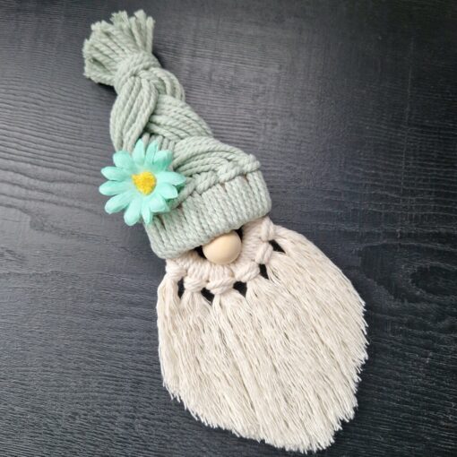 Macrame gnome with braided hat, wooden nose, and white tassel beard, adorned with a green flower, on a dark surface.