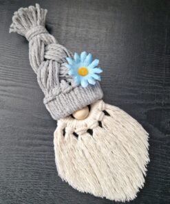 Macrame gnome with a gray braided hat, blue flower, and white fringed beard on a dark surface.