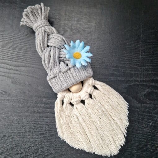 Macrame gnome with a gray braided hat, blue flower, and white fringed beard on a dark surface.