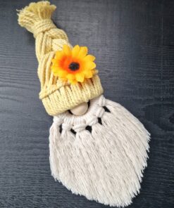 Macramé gnome with yellow braided hat, adorned with an orange flower, and white fringed beard, on a dark surface.