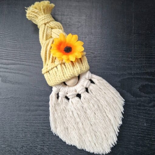 Macramé gnome with yellow braided hat, adorned with an orange flower, and white fringed beard, on a dark surface.