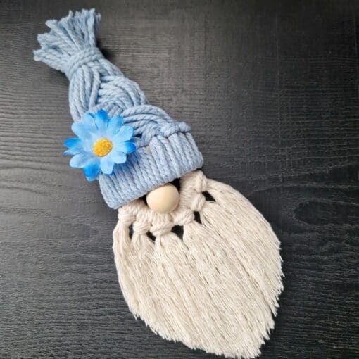 Macrame gnome with a blue hat, decorated with a blue flower, and a long white beard, on a dark wooden surface.