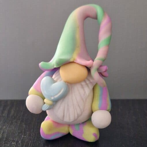 A small gnome figurine with a pastel-colored outfit and hat, holding a blue heart-shaped object, standing on a dark surface.