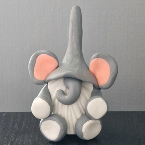 Small clay statue of a gray elephant with large pink ears and a curled trunk, sitting on a black surface against a plain background.