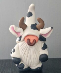 Clay figurine resembling a gnome dressed in a cow costume with horns, black spots, and pink ears on a dark surface.