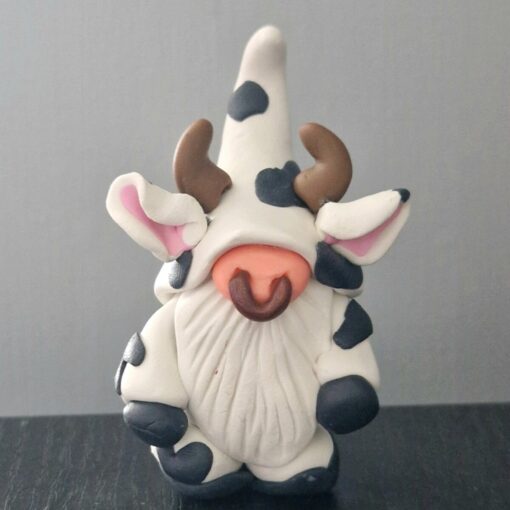 Clay figurine resembling a gnome dressed in a cow costume with horns, black spots, and pink ears on a dark surface.