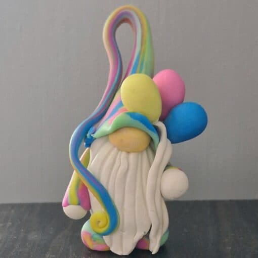 Rainbow clay gnome figure with a white beard, holding colorful balloons, on a gray background.