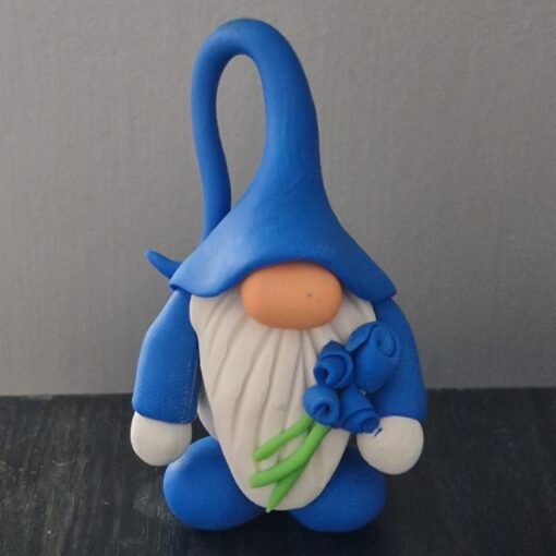 Clay gnome figurine with a blue hat and robe, white beard, holding blue flowers with green stems, set against a gray background.