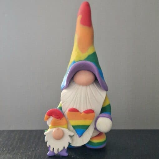 Two gnome figurines in rainbow attire holding a heart with the same colors, set against a plain background.