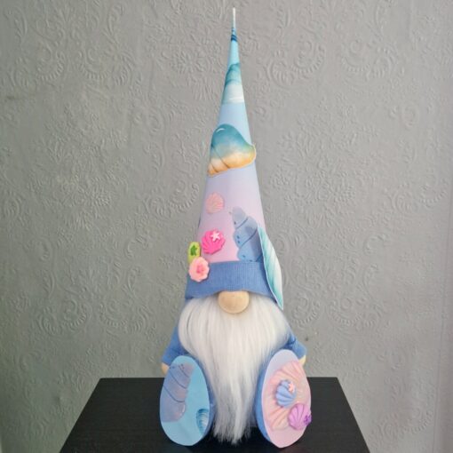 A gnome doll with a long white beard, wearing a tall, pastel-colored hat decorated with seashells, sits against a patterned background.