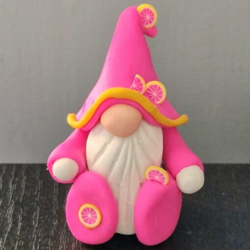 Clay gnome figure with a pink hat and robe, adorned with lemon slices, sitting on a dark surface.