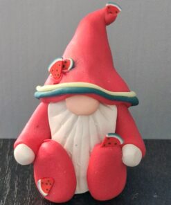 Clay gnome figurine in a red hat and outfit, adorned with small watermelon slice decorations, sitting against a gray background.