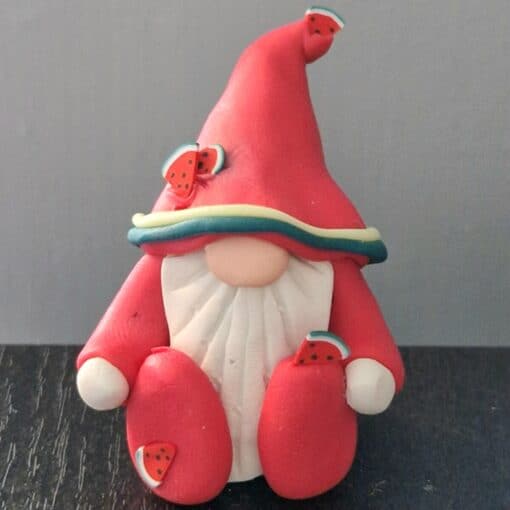 Clay gnome figurine in a red hat and outfit, adorned with small watermelon slice decorations, sitting against a gray background.