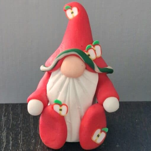 Clay gnome figurine with a red hat, white beard, and apple decorations, sitting on a dark surface with a gray background.