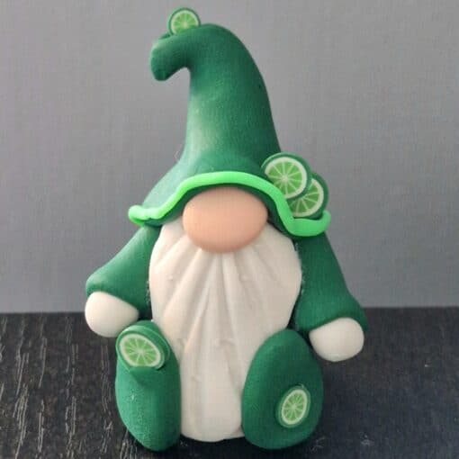 Clay figurine of a gnome with a white beard, wearing a green hat and coat decorated with lime slices, on a dark surface.