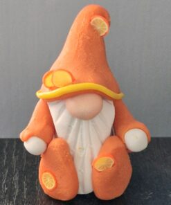 A gnome figurine dressed in orange and white with lemon slice decorations, sitting on a dark surface against a grey background.