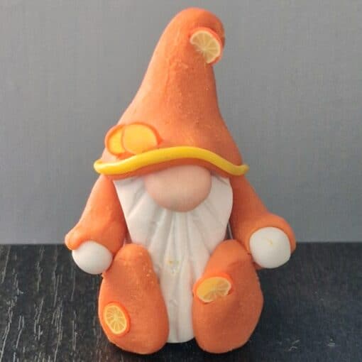 A gnome figurine dressed in orange and white with lemon slice decorations, sitting on a dark surface against a grey background.