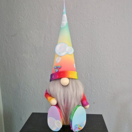 Colorful gnome with a long beard, wearing a rainbow hat decorated with bubbles, sits against a textured, light gray wall.