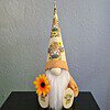 Gnome figure with long white beard and tall hat featuring cow images, holding a sunflower, sits against a textured gray background.