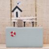 Miniature wooden beach hut model with stairs, a lifebuoy, and a patterned flag on top against a rustic wooden background.