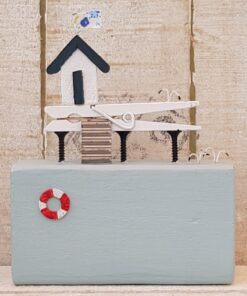 Miniature wooden beach hut model with stairs, a lifebuoy, and a patterned flag on top against a rustic wooden background.